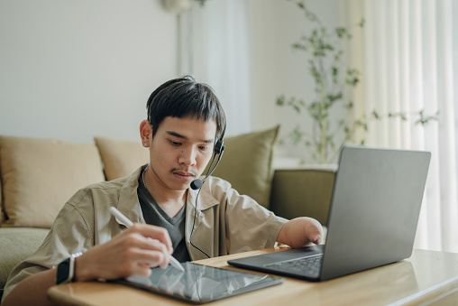 Asian man with arm disabilities sitting at home and working on laptop and doing video communication with his colleague.