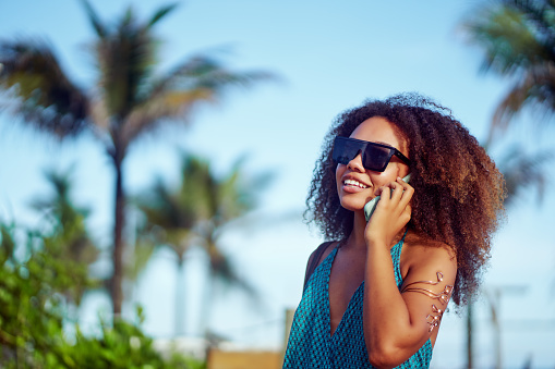 young brazilian black girl with afro hairstyle talking on the cell phone on the beach with palm trees and sky in the background in brazil