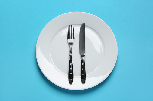 Clean plate with shiny silver cutlery on light blue background, top view