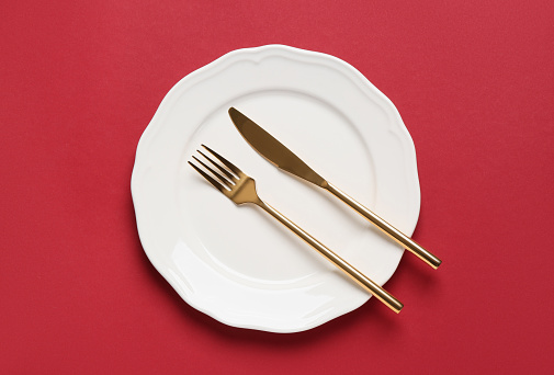 Clean plate with golden cutlery on red background, top view