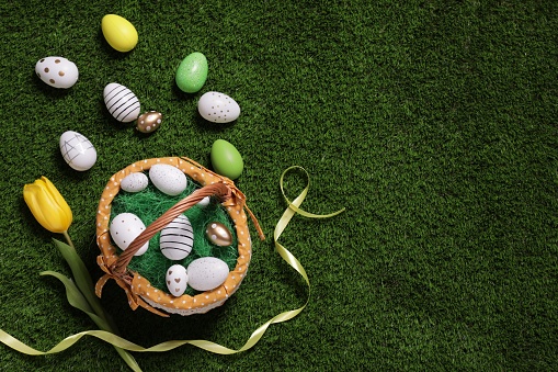 Easter basket with painted eggs, tulip and ribbon on green grass, flat lay. Space for text
