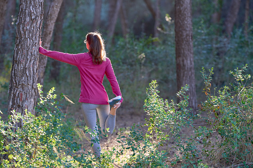 sporty woman stretching her legs leaning on tree in the forest