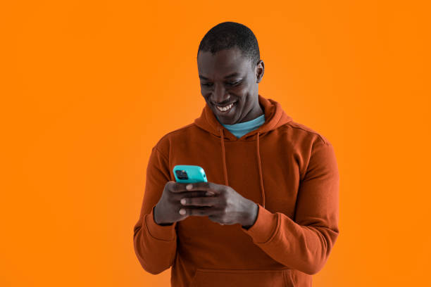 Homens afro-americanos sorridentes usando telefone e cartão de crédito em pé na frente do fundo laranja - foto de acervo