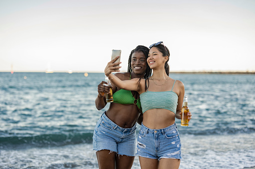 two multiracial female friends on the beach take selfie with smart phone, on the beach at sunset.