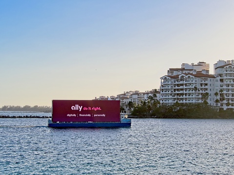 Miami Beach, FL - USA, February 1, 2023. A floating digital billboard reenters the port of Miami after advertising along the Miami beach shoreline. A Ballyhoo Media vessel shown floating by Miami high end condominiums as it enters Miami harbor near sunset. An Ally bank advertisement shown with copy space.