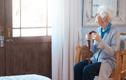 Thinking, sad and senior woman thinking of life with a walking stick by the window in the room in a retirement house. Elderly female with depression, disability and retirement idea in a nursing home