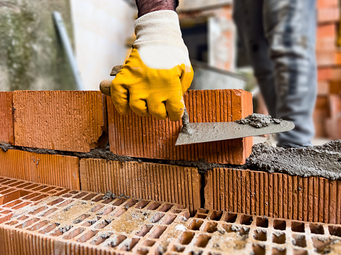 Masonry craftmenship concept: Close-up of industrial bricklayer installing bricks on construction site. Professional worker building exterior walls, using protective gloves and trowel for laying cement between bricks. Detail of mason with tools and concrete.