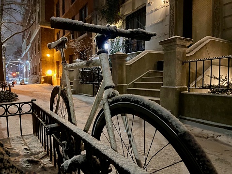 Close-up of a white bicycle with its frame and wheel chained to a metal tree guard on a New York City sidewalk during a snow storm at night