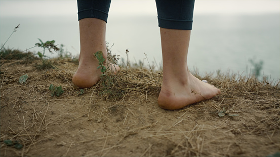 Close up woman feet standing sand hilltop in front grey ocean. Unknown barefoot girl stop on dry beach grass relaxing outdoors. View of sporty legs wearing black leggings on cloudy summer nature.