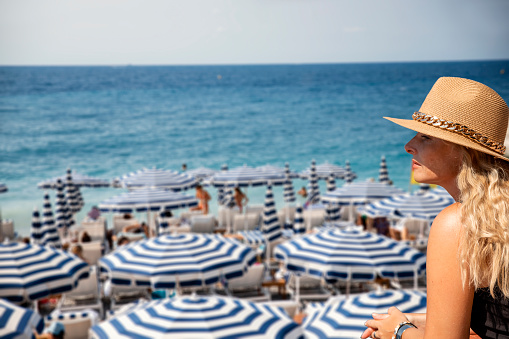 Side view of elegant blonde woman with sun hat looking at blue-white parasols on French Riviera sea beach. Copy space