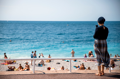 Back of Arabian Woman with headscarf using phone to take a photo of French beach at Nice waterfront. Copy space