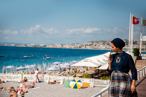 Arabian Woman with headscarf walking at Nice waterfront. Thoughtful female with phone on mouth looking for someone at city beach. Copy space