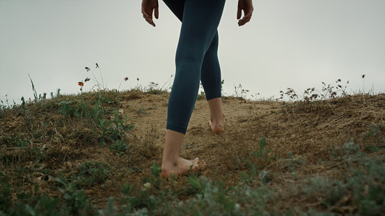 Barefoot sporty girl climbing green hill wearing black leggings close up. Athletic woman legs stop on sandy hilltop gloomy morning. Young lady feet walking seacoast cloudy day. Nature concept.