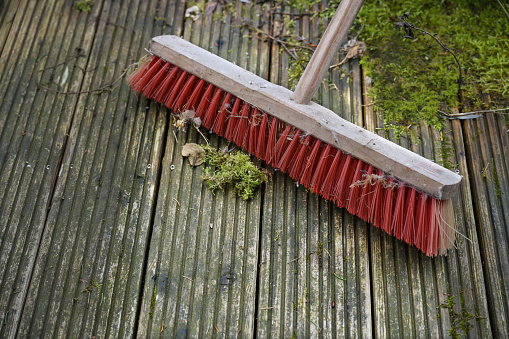 Red outdoor broom with stiff plastic bristles on a dirty wooden deck with algae and moss, spring cleaning in garden and yard for the new season, copy space, selected focus, narrow depth of field