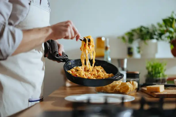 An anonymous chef in apron picking up tagliatelle with serving tongs from the frying pan and serving it on the plate.