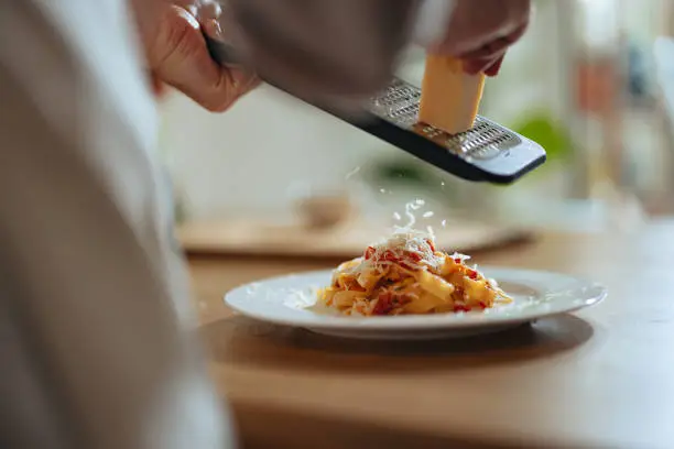 Unrecognizable chef in apron grating parmesan cheese in homemade pasta in the kitchen.