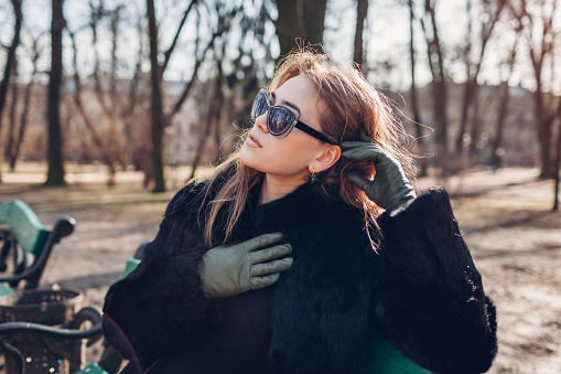 Close up portrait of stylish young woman wearing fur coat, green leather gloves and sunglasses in park. Spring trendy accessories. Girl relaxing in park