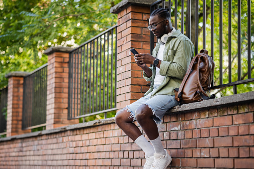 A modern young man is using a mobile phone and enjoying city life. He is scrolling through his social media feed while sitting on the brick wall. Lifestyle and technology.