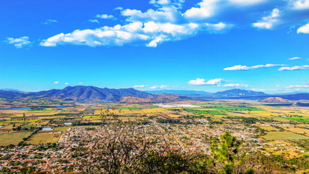 The city of Etzatland Jalisco Mexico as seen from above.  The city is sprawled out and there are distant agricultural fields and a mountain range.  This is a wide angle panoramic shot with lots of open copy space. A beautiful blue sky is punctuated by white fluffy clouds.