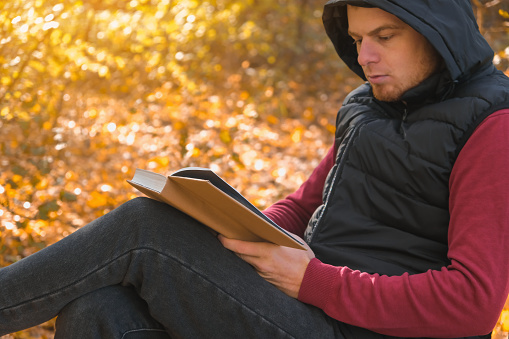 Young man is sitting on chair in an autumn park or forest and reading book. Mental recreation in nature. Quiet country life. Escapism. Digital detoxification. Concept of reading book in an autumn park