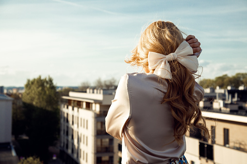 Pretty girl in romantic outfit with wonderful hairstyle and big elegant bow standing back and looking around from observation platform closeup. Trend look, relaxation on roof, sky, paris cityscape.