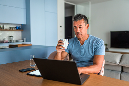Front View Of A Mature Japanese Man Working From Home Using Laptop And Drinking A Cup Of Coffee