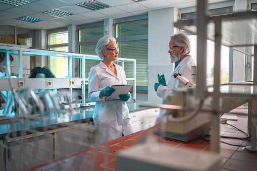 Caucasian female and male  experienced professionals leaning on the lab counters, wearing white uniforms,  gloves and protective eyewear, talking face to face. Side view of both, partly shot through blurred laboratory equipment. Younger colleagues in background.