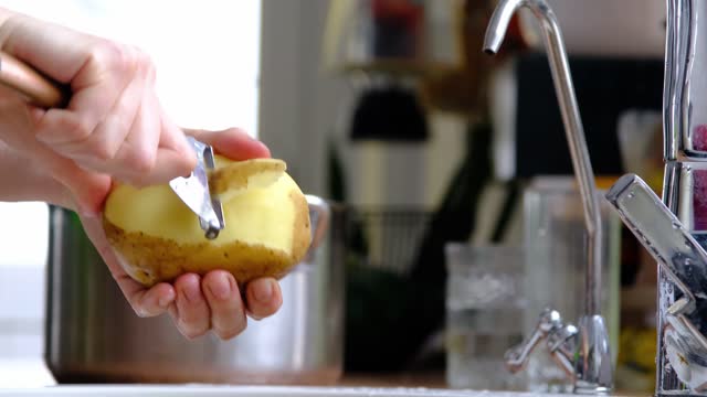 Women's hands peel raw potatoes with a special knife with a vegetable peeler under a stream of water in the kitchen. Cooking, saving water, eco-friendly use of resources