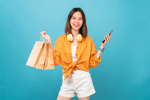Beautiful Asian woman holding brown blank craft paper shopping bags and showing smartphone on blue background.