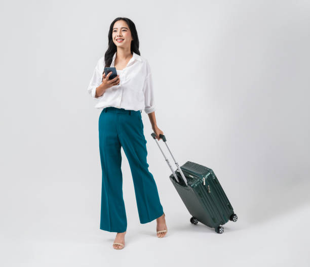 Cheerful young Asian woman using smartphone and holding suitcase with walking to the front, isolated on white background. Concept tourist holiday trip. stock photo