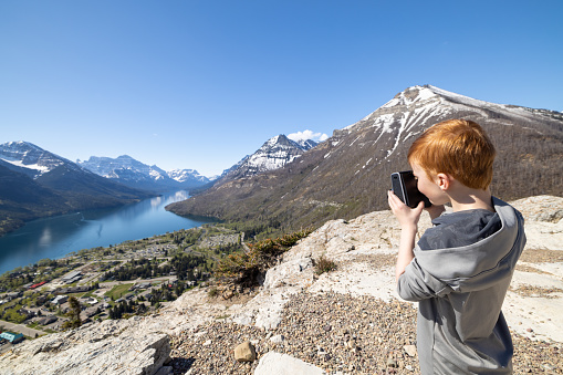 Young Redhead Boy Taking Instant Picture at the top of Bear's Hump Trail in Waterton Lakes National Park, Alberta, Canada on a nice day of summer. He is looking at the picture after the shot.