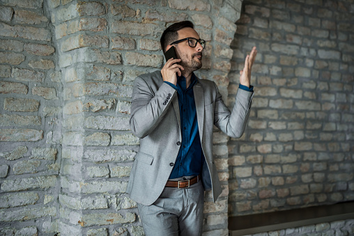 Frustrated young entrepreneur communicating over cell phone while leaning on a brick wall of his office.