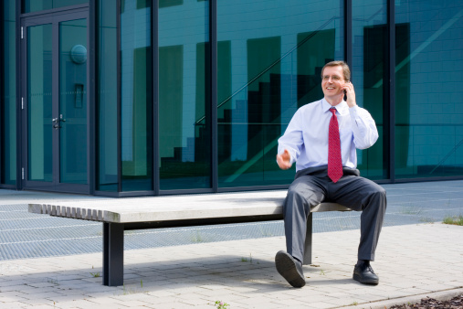 Businessman sitting on a bench in front of an office building and talking on mobile phone
