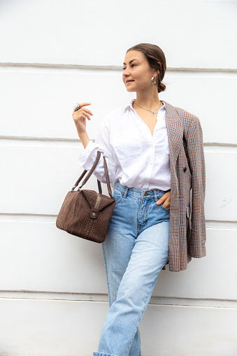Stylish slim woman in white shirt and oversize jacket with brown bag against wall in street, fashion trend, urban casual style