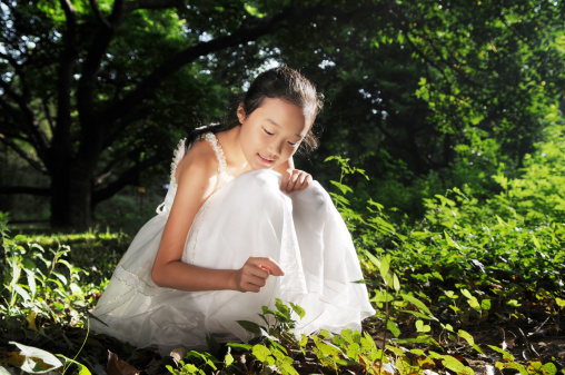 A smiling Chinese girl in white gauze skirt squatting on the meadow in grove in summer morning sunshine.
