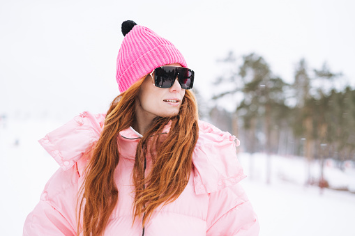 Young red haired woman in pink sportswear and sunglasses on winter snowy background