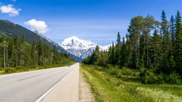 Road leads to to Mount Robson with slightly clouded blue sky and highway and green gorest on foreground
