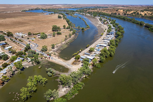 Hope after the flood: aerial view of houses, shacks drying out as Murray River floodwaters recede. Vehicles line the roadside as owners return to begin the cleaning up process with some some properties still surrounded by water and the main access road still closed. High water marks can be seen on sheds & water tanks. A speedboat creates a wake in the main river as it slows to stop.  Bow Hill, South Australia
