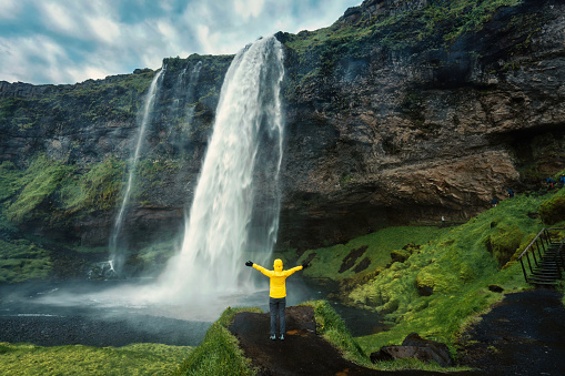 Cheerful asian woman in yellow jacket standing and looking at moody Seljalandsfoss waterfall flowing from cliff in summer at South of Iceland