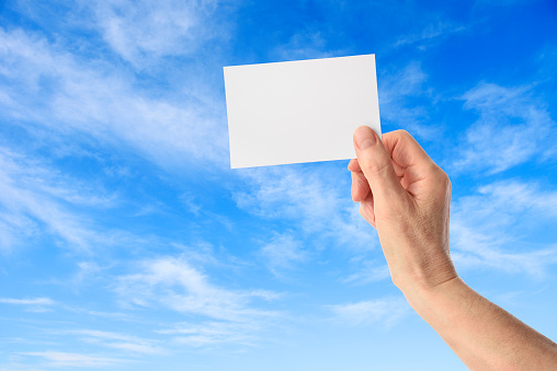 Close-op of hand holding a blank paper against blue sky.