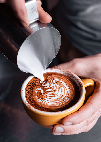 A barrister's hands are seen pouring frothed milk into a large coffee cup, forming a squiggly pattern on the surface of the coffee.