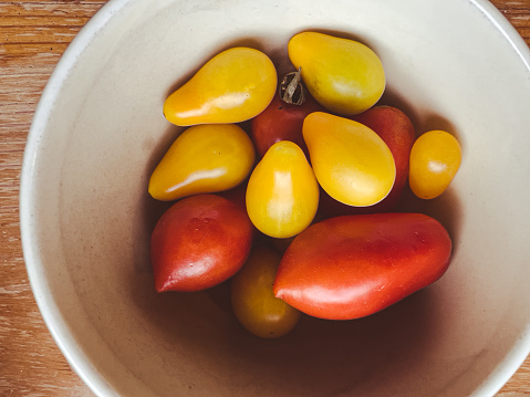 Horizontal high angle closeup photo of tiny red and yellow pear shaped organic tomatoes in a round white ceramic bowl on a rustic wooden table.