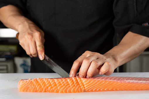A chef's hands are seen slicing up a large piece of sashimi grade salmon in a restaurant kitchen.