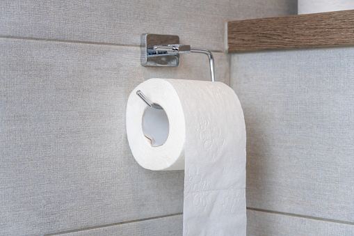 Roll of white toilet paper on a shiny holder hanging on a light gray tile wall. Copy space