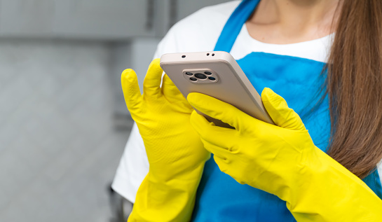 Close-up of a woman in yellow rubber gloves and a blue apron holding a smartphone and reading the news in the kitchen interior. Application and blog for social networks, house cleaning