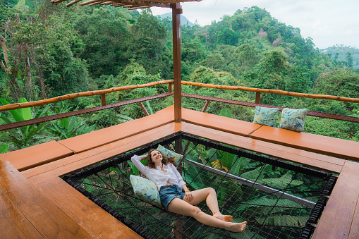 Young Caucasian woman in positive mood  laying in hammock on the terrace and looking at the jungles