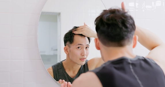 asian man standing in front of mirror is concerned about hair loss or alopecia