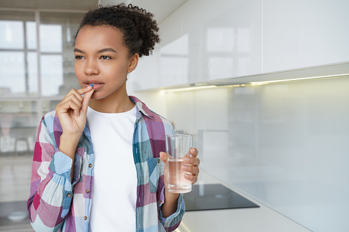 Pensive mixed race girl takes a pill holding glass of water, standing at home. Unhealthy young female taking medicine tablet, antibiotic, painkiller or vitamins, sedatives. Health care, flu cure.