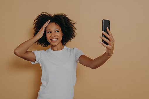 Happy charming dark skinned female 20s in white tshirt making photo selfie on modern smartphone and smiling cheerfuly, isolated over beige wall. Positive young african woman taking photo on cellphone