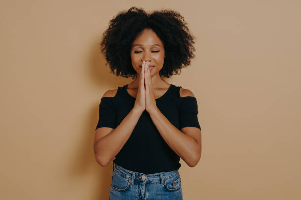 jeune femme afro-américaine priant et se tenant la main dans le geste de prière debout dans le studio - black celebration colors dark photos et images de collection